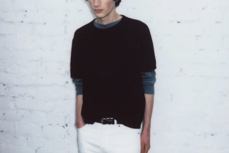 a model wearing a black shirt and white pants against a white concrete wall.