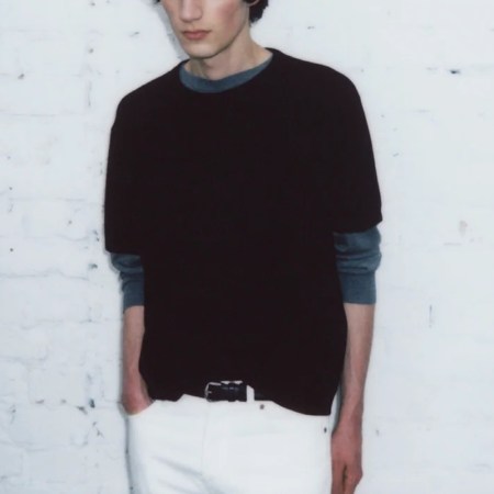 a model wearing a black shirt and white pants against a white concrete wall.