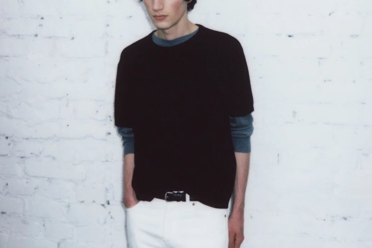 a model wearing a black shirt and white pants against a white concrete wall.