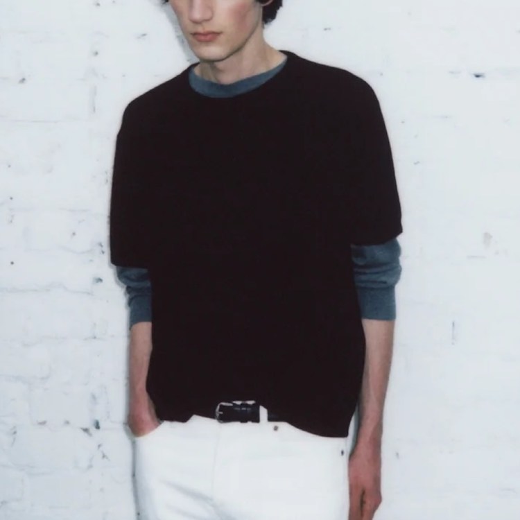 a model wearing a black shirt and white pants against a white concrete wall.