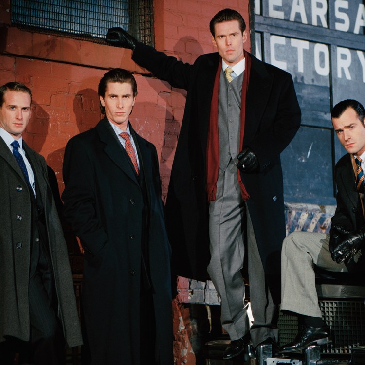 Four men in dark overcoats and suits pose against a brick wall and car, one holding a cigarette.
