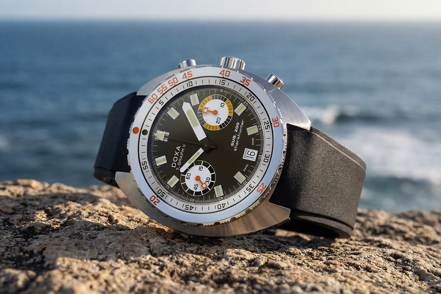 A dive watch with a black dial and black strap sitting on a rock with the ocean in the background