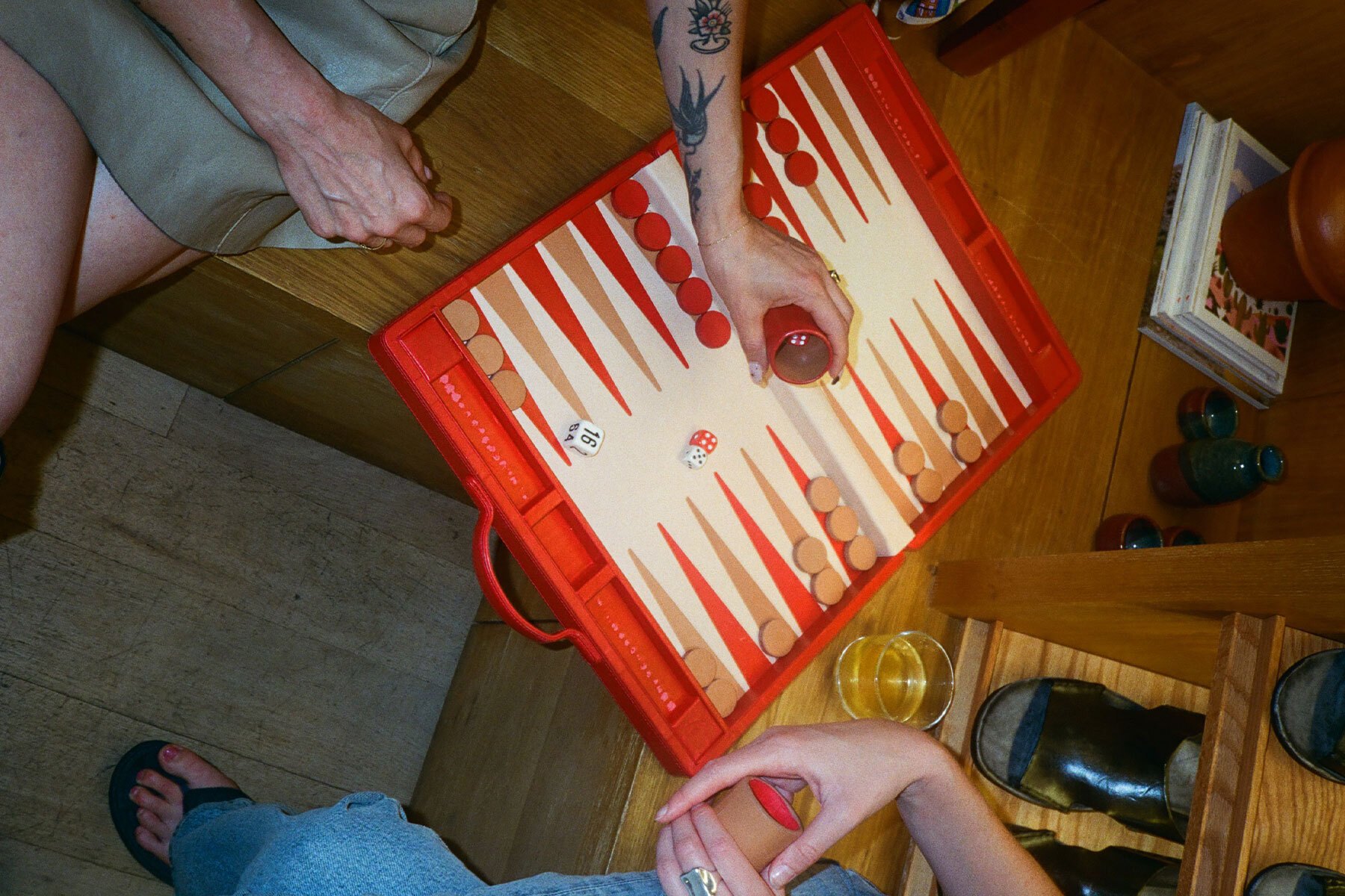 A red backgammon board with hands holding dice cups hovering over it as two people play the game
