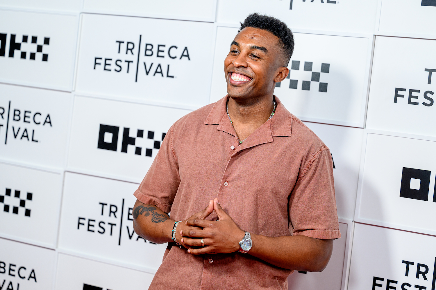 "Survivor" champion Kyle Fraser smiling in a rust-colored shirt with a beaded necklace and watch, hands clasped, against a Tribeca Festival backdrop.
