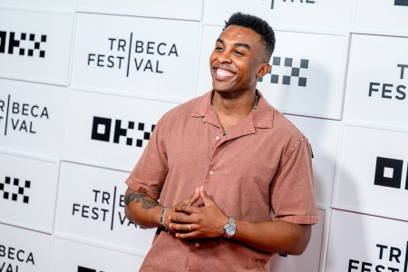 "Survivor" champion Kyle Fraser smiling in a rust-colored shirt with a beaded necklace and watch, hands clasped, against a Tribeca Festival backdrop.