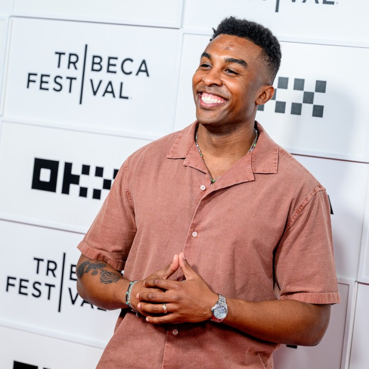 "Survivor" champion Kyle Fraser smiling in a rust-colored shirt with a beaded necklace and watch, hands clasped, against a Tribeca Festival backdrop.