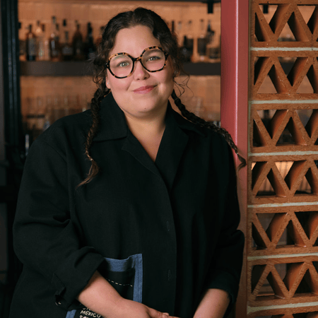 A smiling woman with braided hair and tortoiseshell glasses stands in a restaurant next to a decorative brick wall.