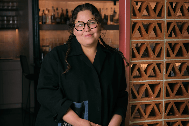 A smiling woman with braided hair and tortoiseshell glasses stands in a restaurant next to a decorative brick wall.