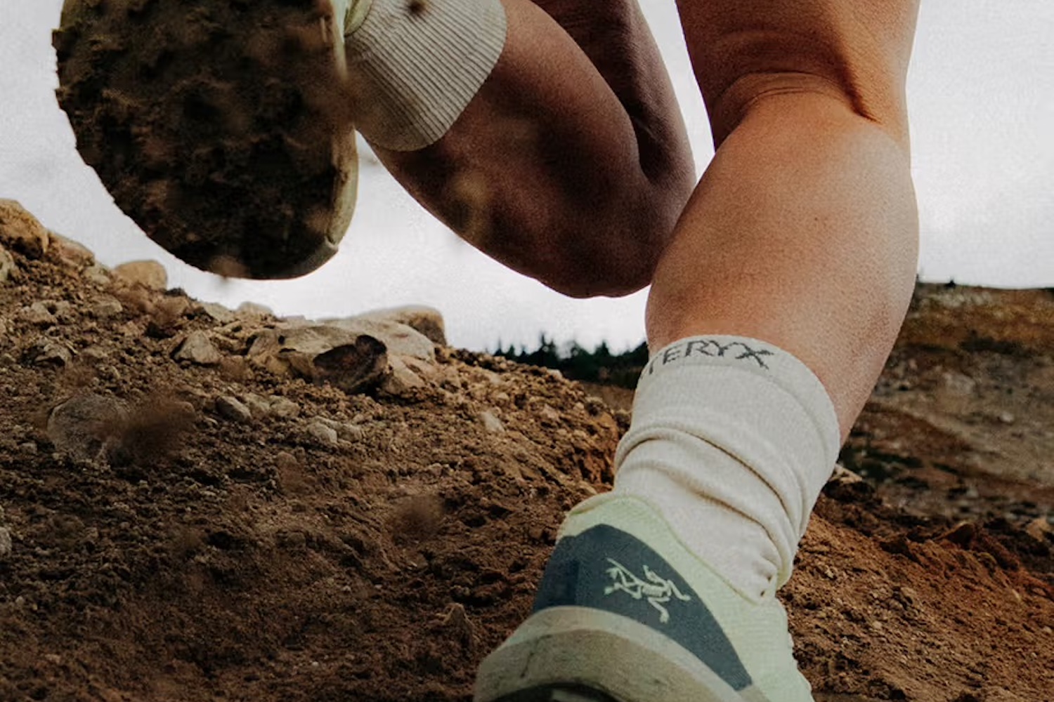 Close-up of a person's legs and feet in trail running shoes and socks, kicking up dirt on a rocky path.