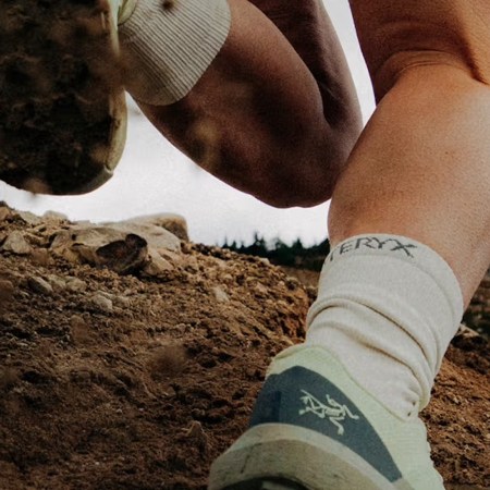 Close-up of a person's legs and feet in trail running shoes and socks, kicking up dirt on a rocky path.