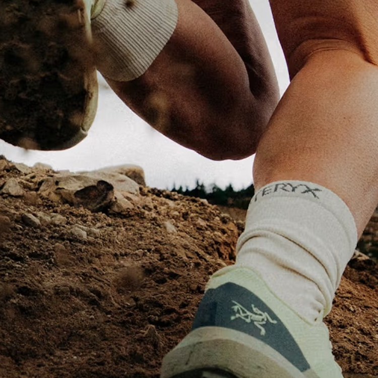 Close-up of a person's legs and feet in trail running shoes and socks, kicking up dirt on a rocky path.