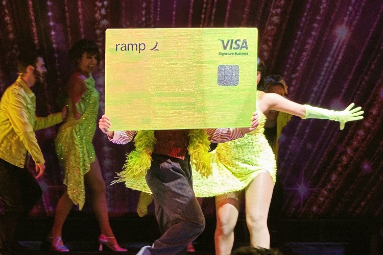 Performers in bright neon outfits on a stage, holding up a large yellow Ramp Visa card.