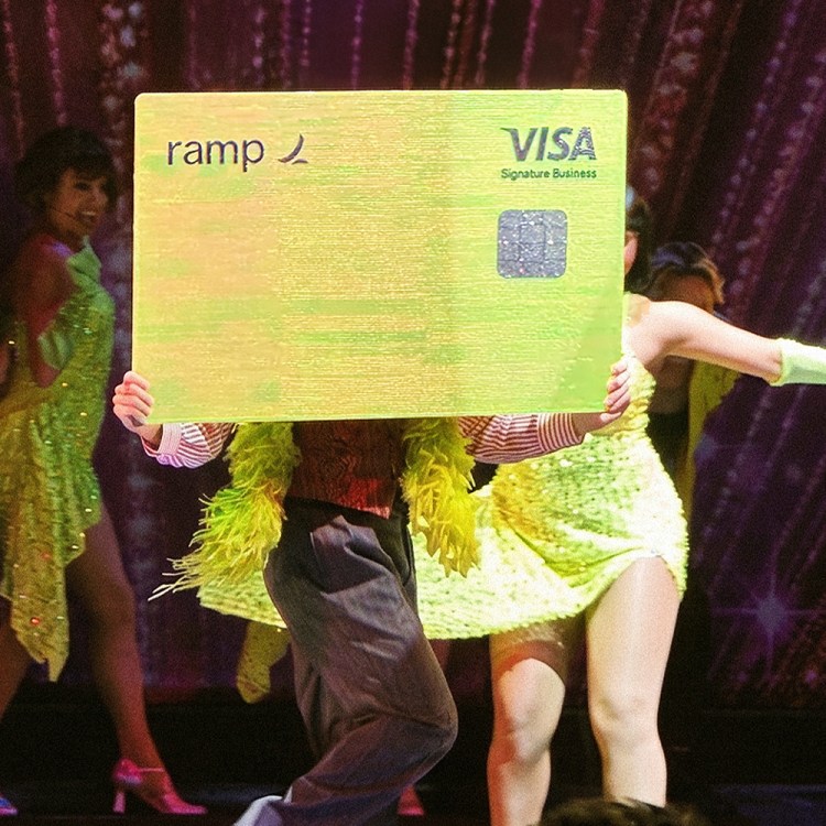 Performers in bright neon outfits on a stage, holding up a large yellow Ramp Visa card.