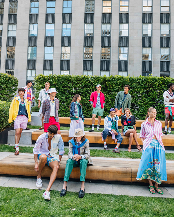 Models in colorful, preppy outfits pose on wooden benches and grass in front of a modern building.