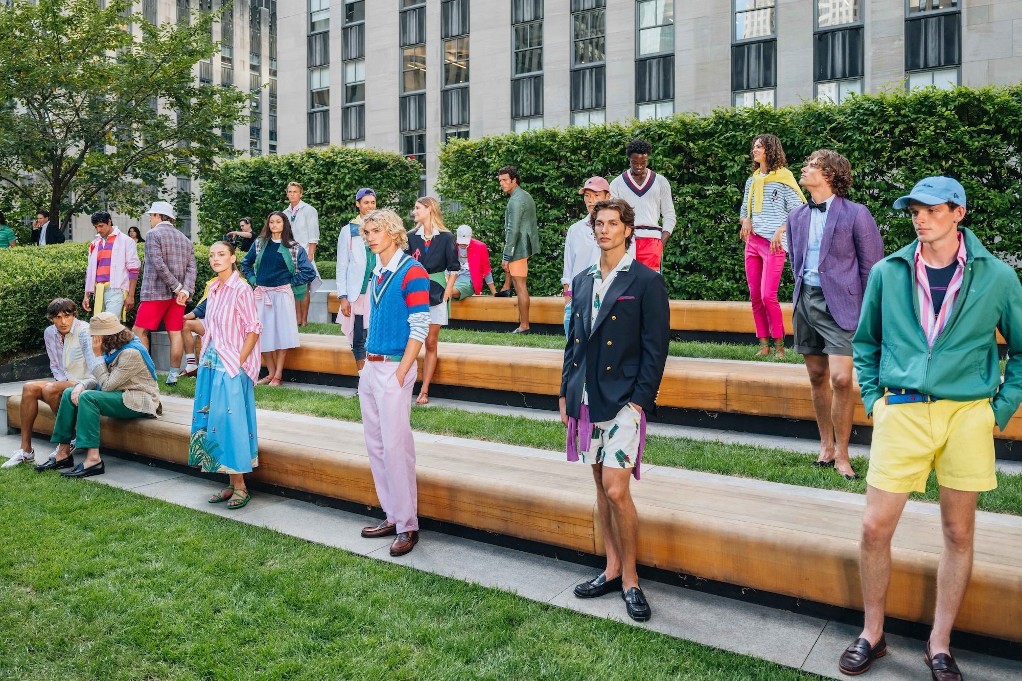 Models in colorful, preppy clothing pose on wooden benches in a garden with a city building backdrop.