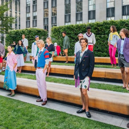 Models in colorful, preppy clothing pose on wooden benches in a garden with a city building backdrop.