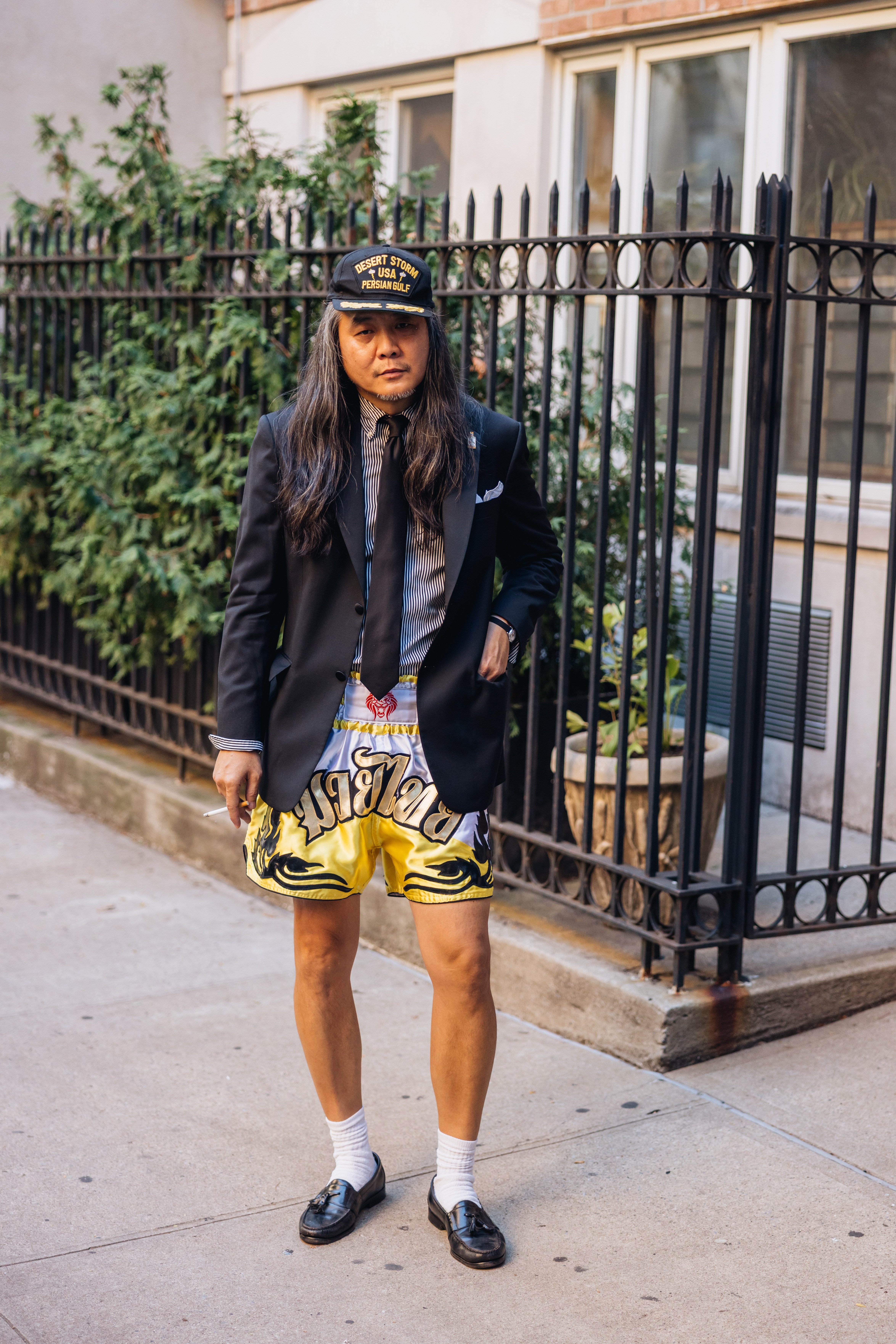 A man with long hair and a "Desert Storm" cap wears a suit jacket, striped shirt, tie, yellow Muay Thai shorts, white socks, and loafers.