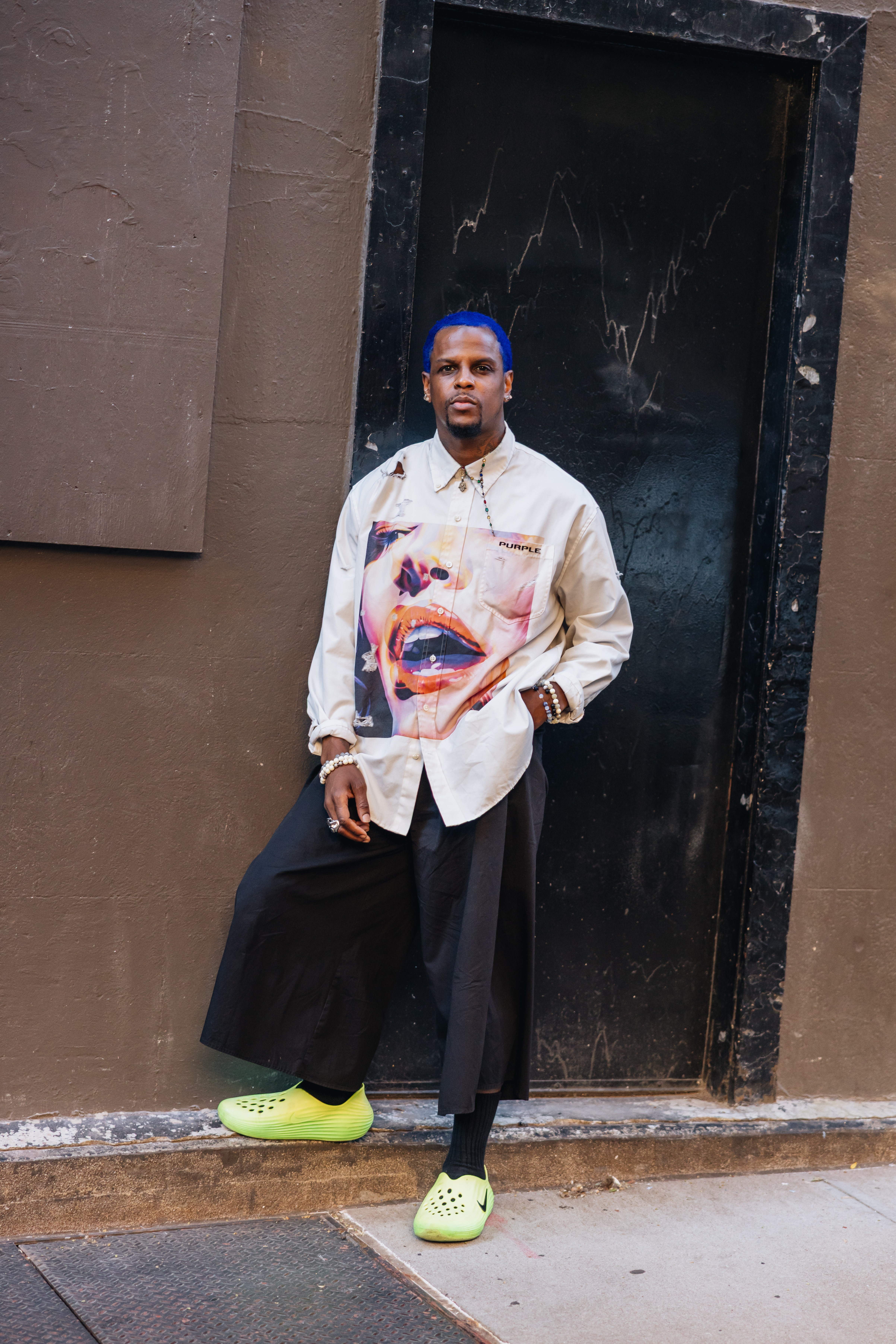 A man with blue hair and a white shirt with a graphic of a woman's face, black wide-leg pants, and neon green shoes.