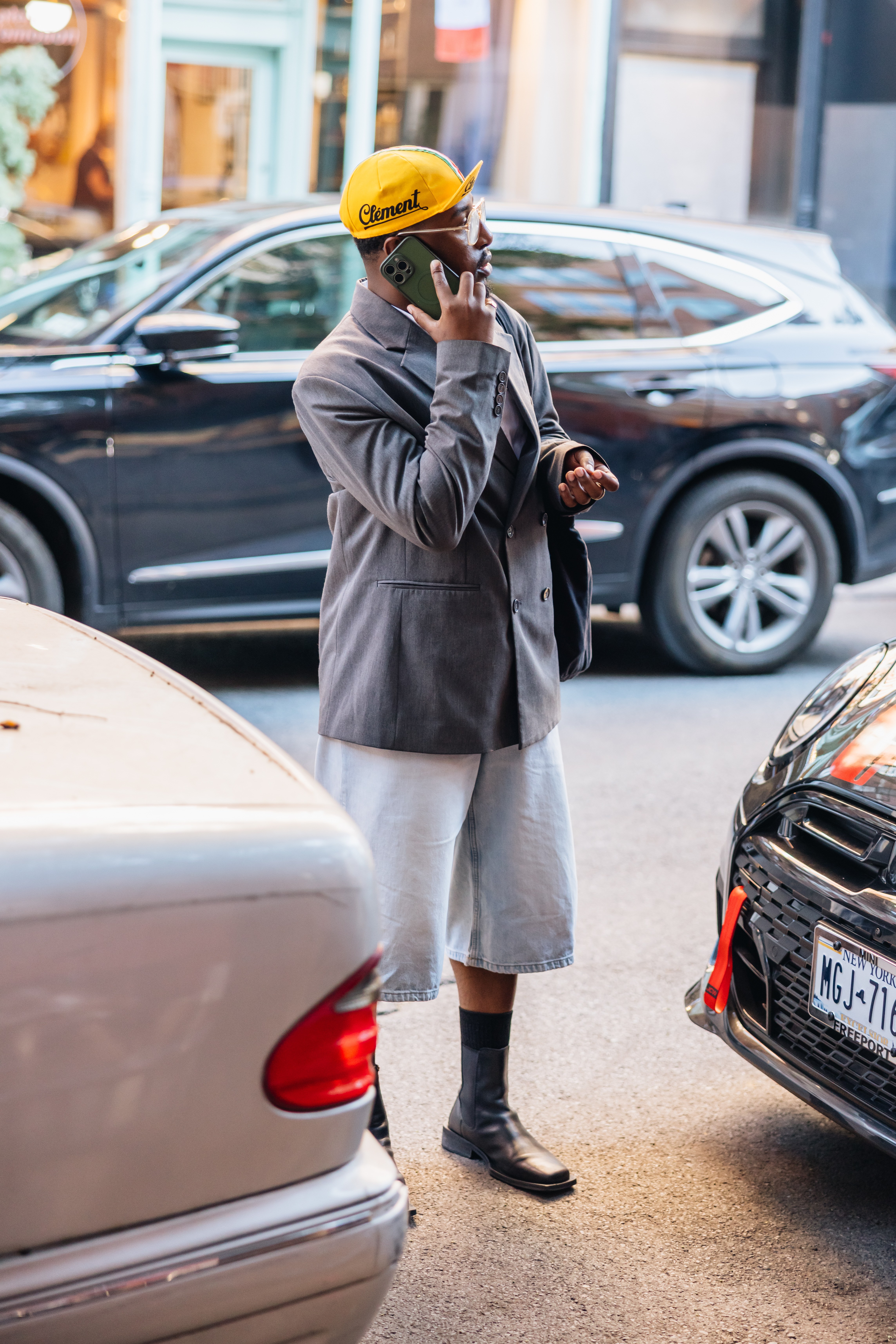 Man in a gray blazer, light wash denim shorts, and black boots talks on a phone, wearing a yellow "Clement" cap.
