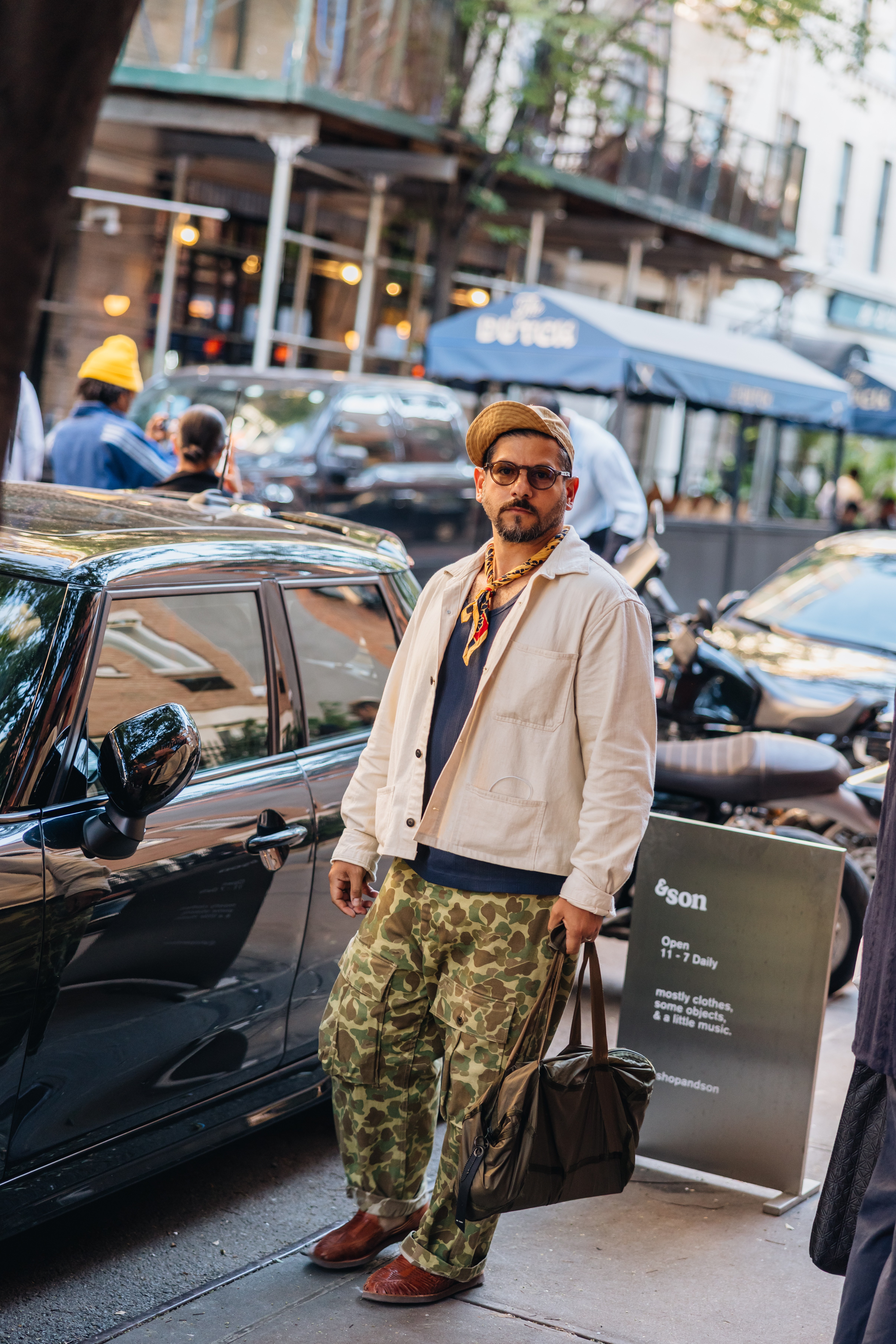 Man in a tan jacket, camo pants, and a flat cap, holding a bag, stands by a black car on a city street.
