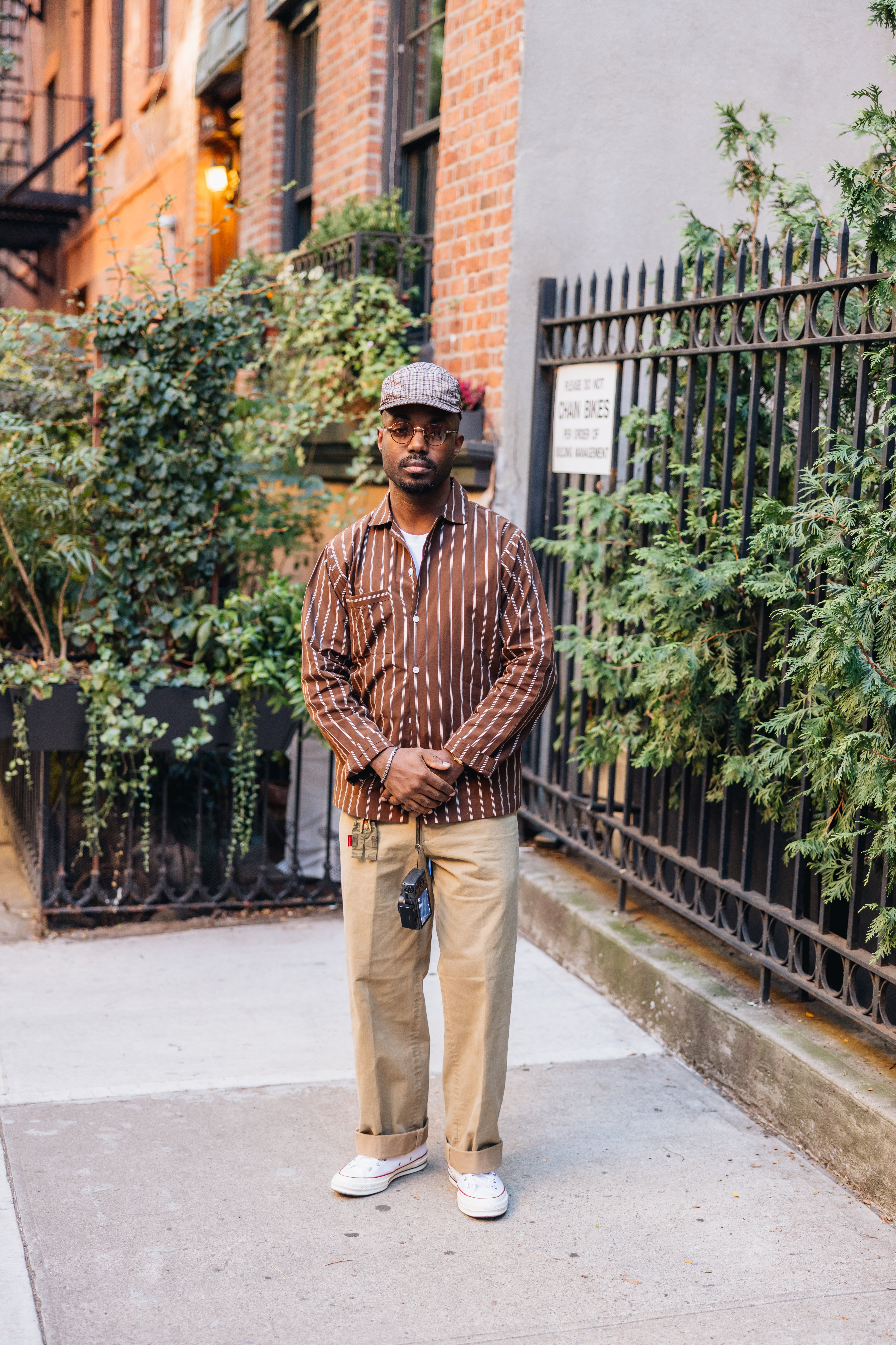 A man in a brown striped shirt, khaki pants, and a plaid cap stands on a city sidewalk, hands clasped.