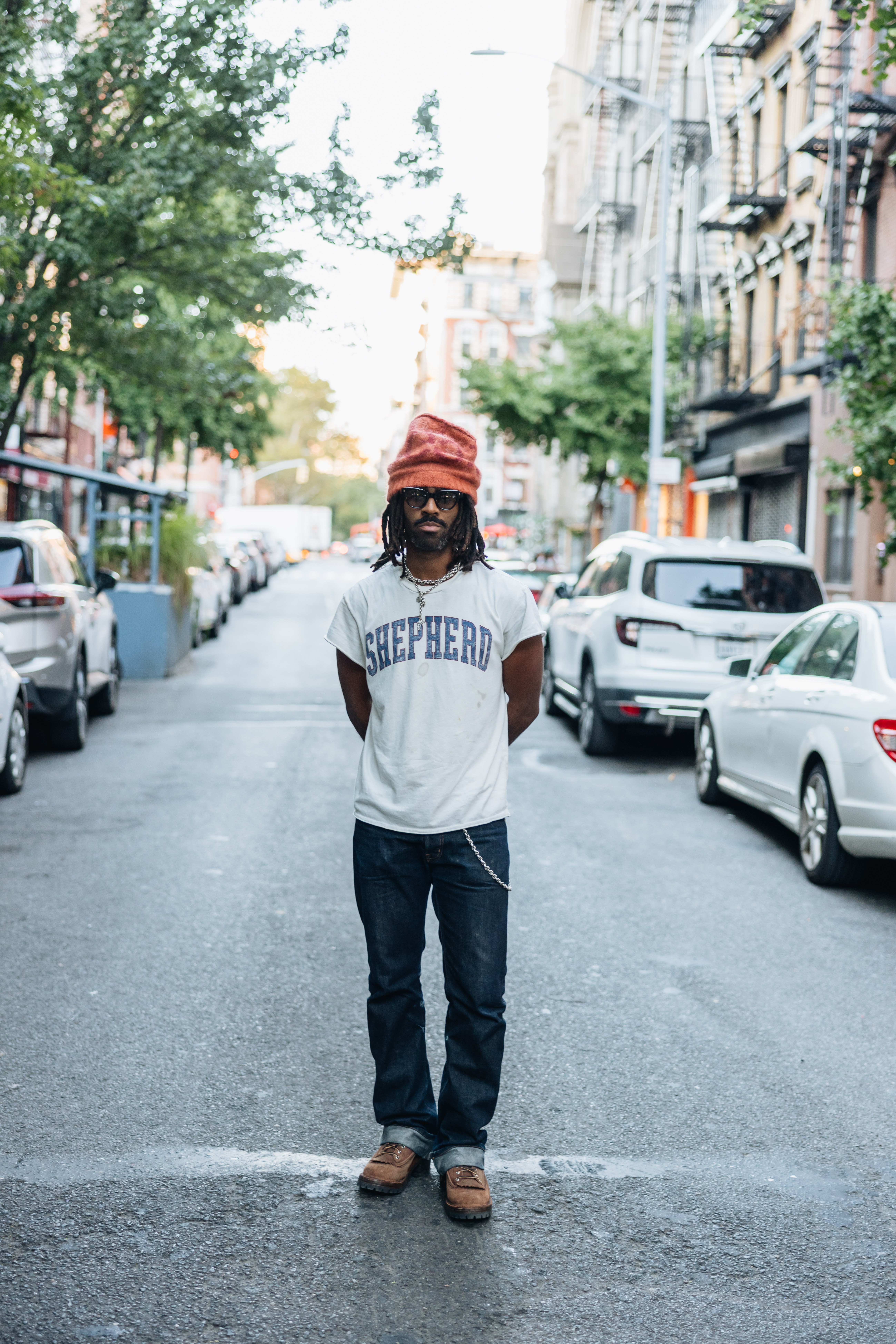 Man in a red beanie, sunglasses, and "SHEPHERD" t-shirt stands in the middle of a city street.