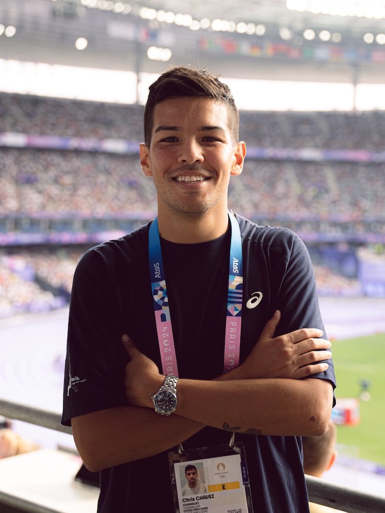 Smiling man with dark hair and a lanyard, arms crossed, in a stadium.