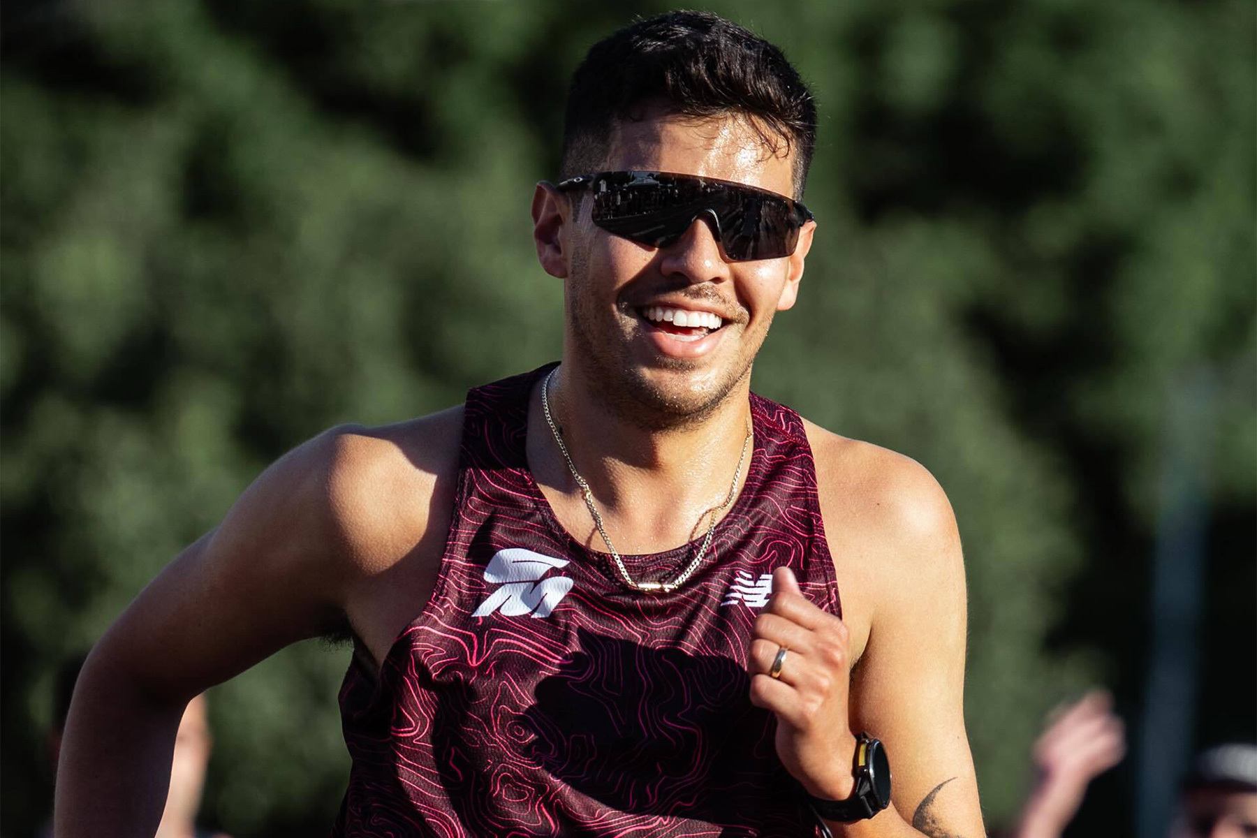 Sweaty man with dark hair, sunglasses, and a maroon tank top with a topographic pattern, smiling while running.