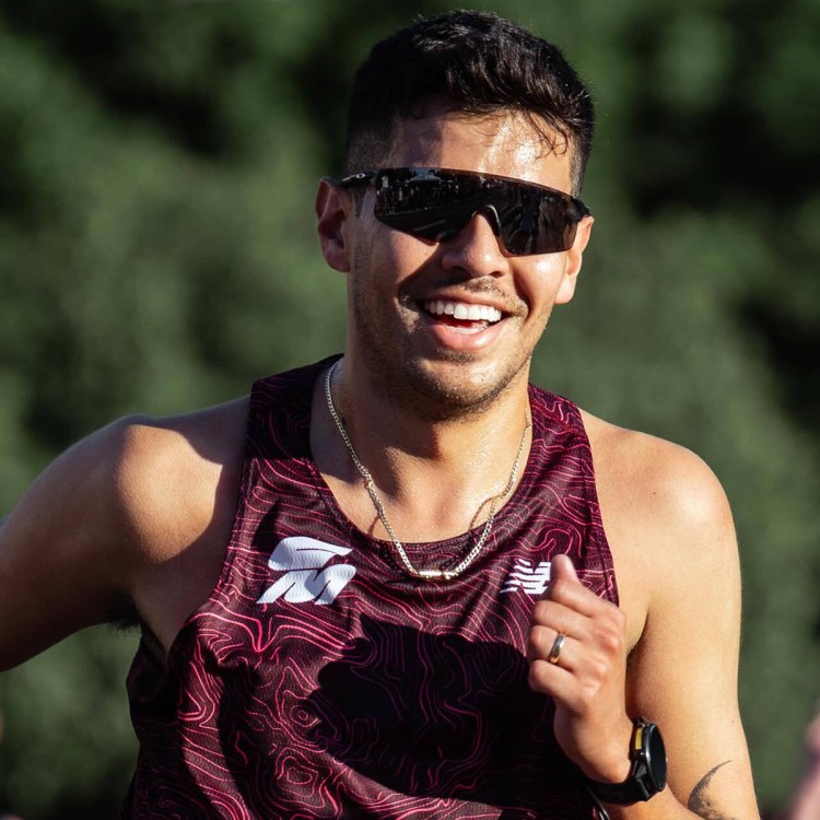 Sweaty man with dark hair, sunglasses, and a maroon tank top with a topographic pattern, smiling while running.