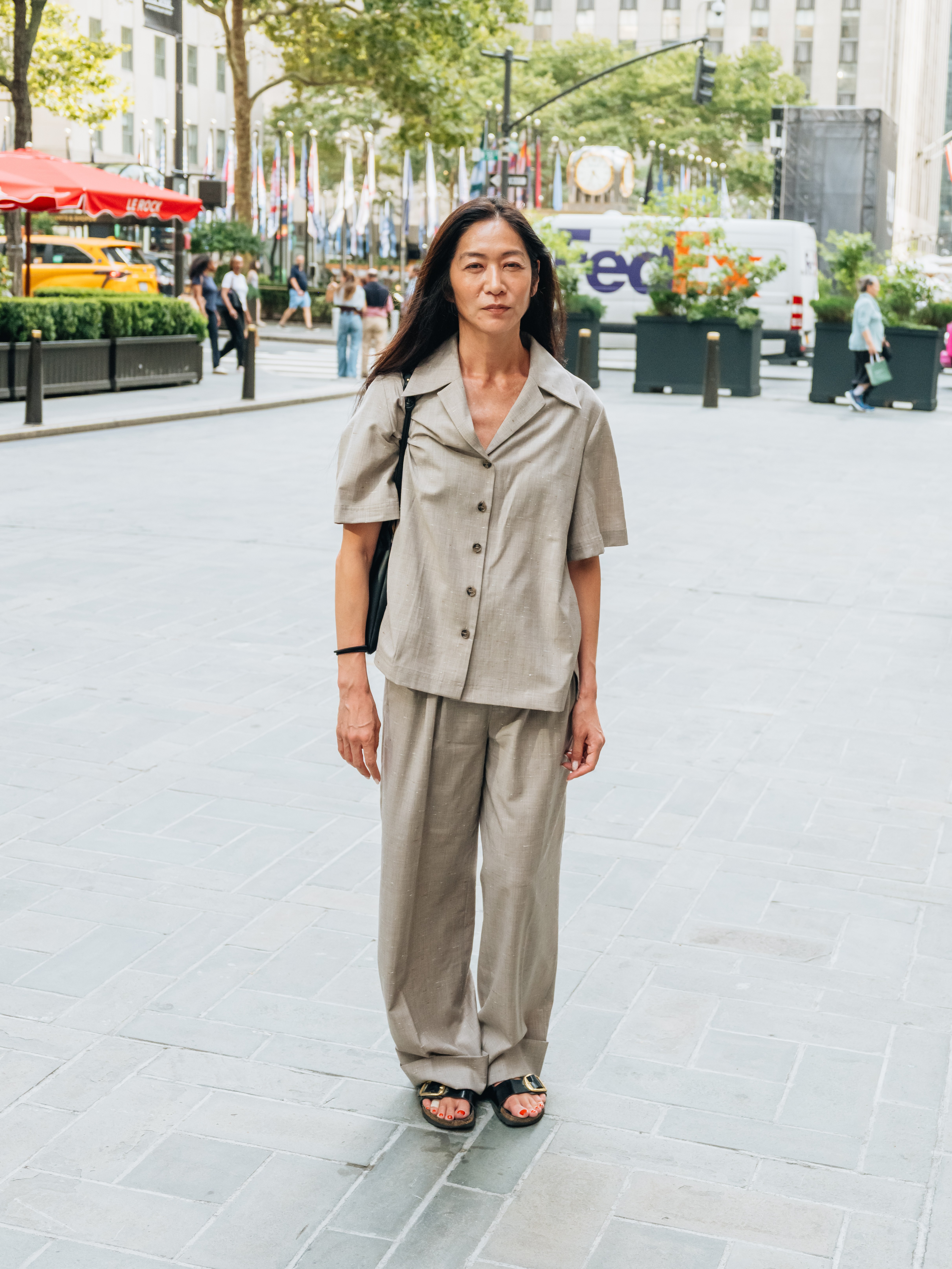 A woman with long dark hair wears a matching light brown short-sleeved button-up and wide-leg pants, with black sandals.