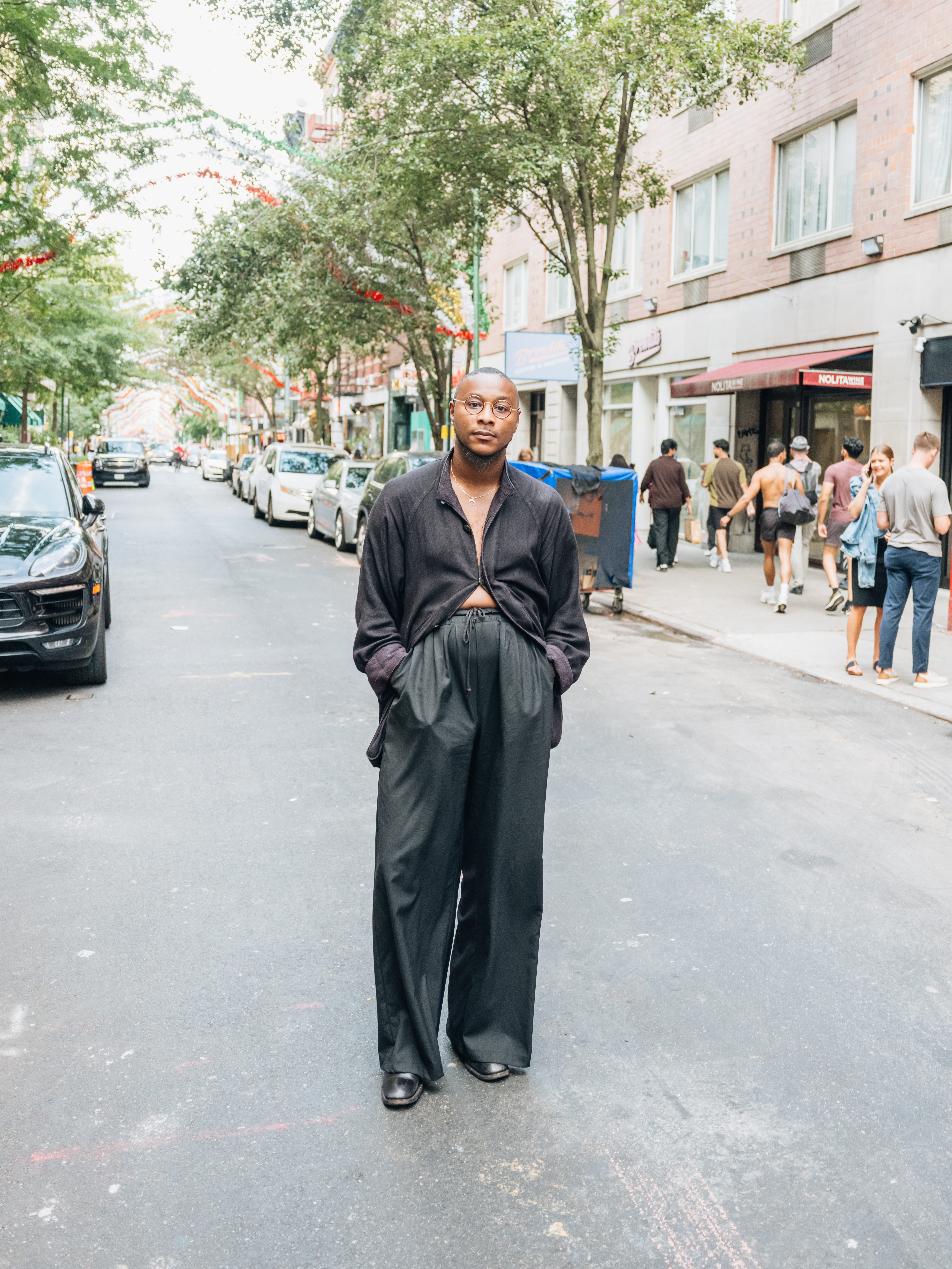 A bald Black man in round glasses, an open black shirt, and wide-leg black pants stands on a city street.