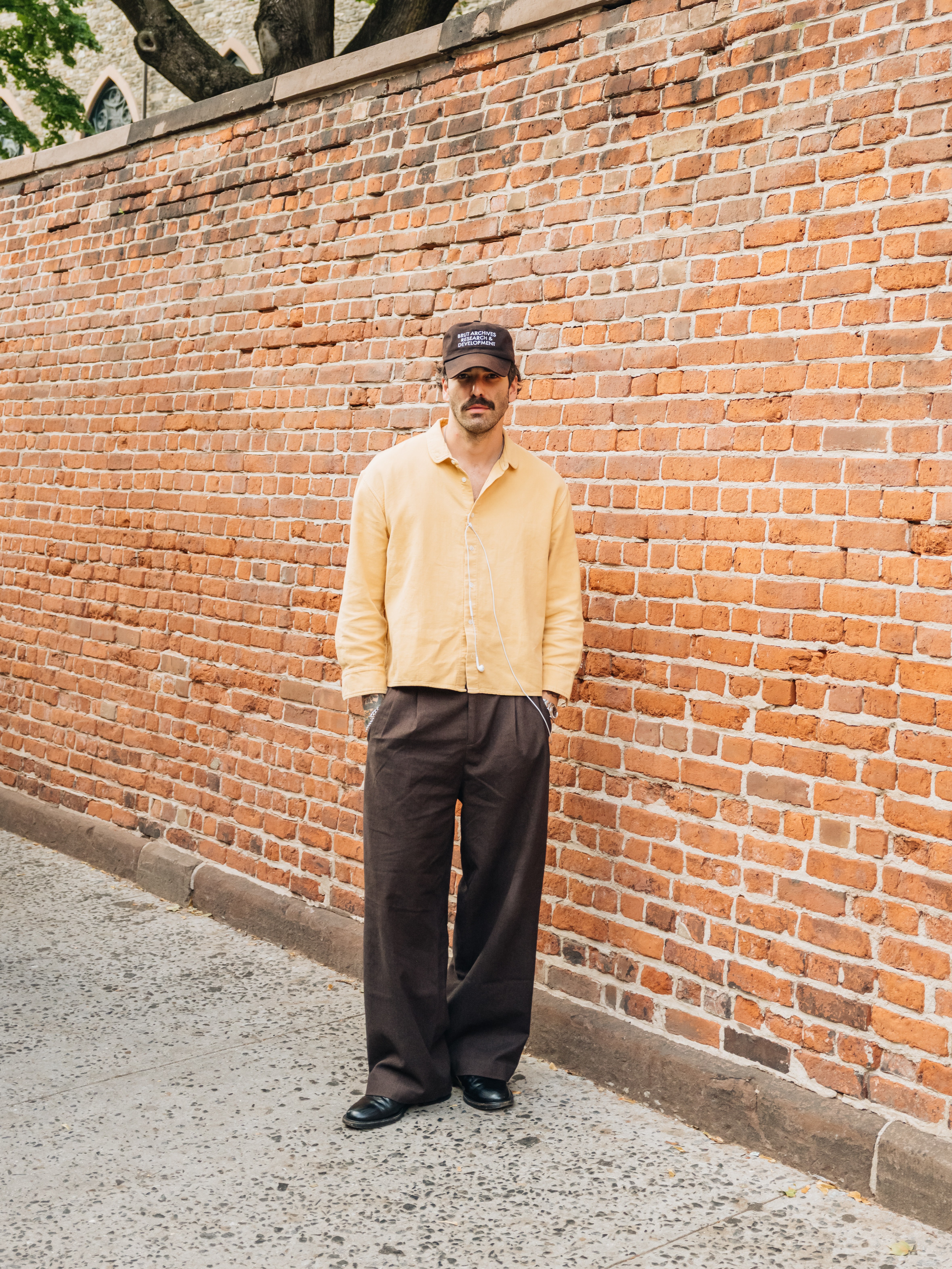 Man in a brown cap, yellow shirt, and dark brown pants stands against a brick wall, hands in pockets.