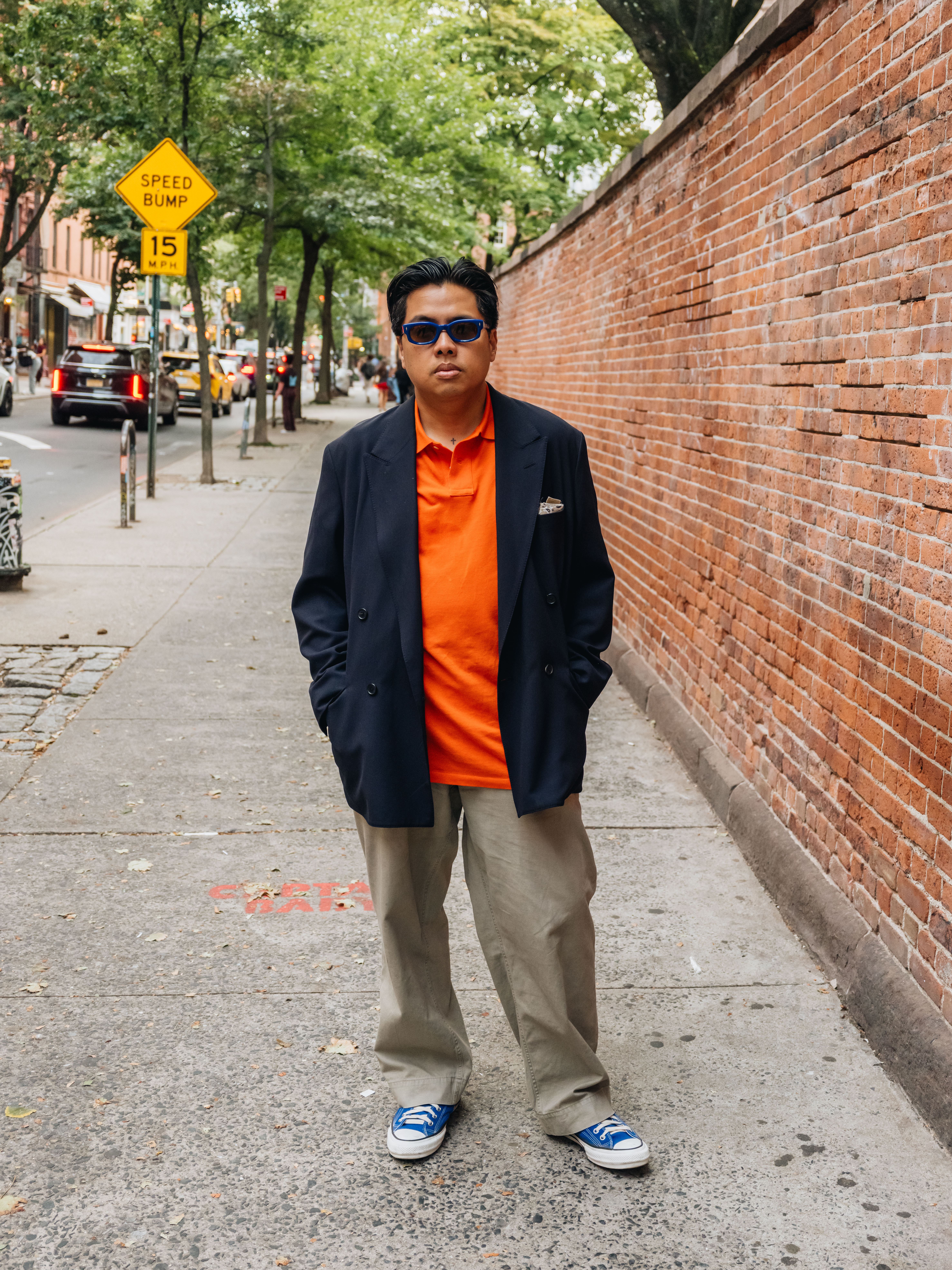 A man in blue sunglasses, an orange polo, navy blazer, khaki pants, and blue sneakers stands on a city sidewalk.