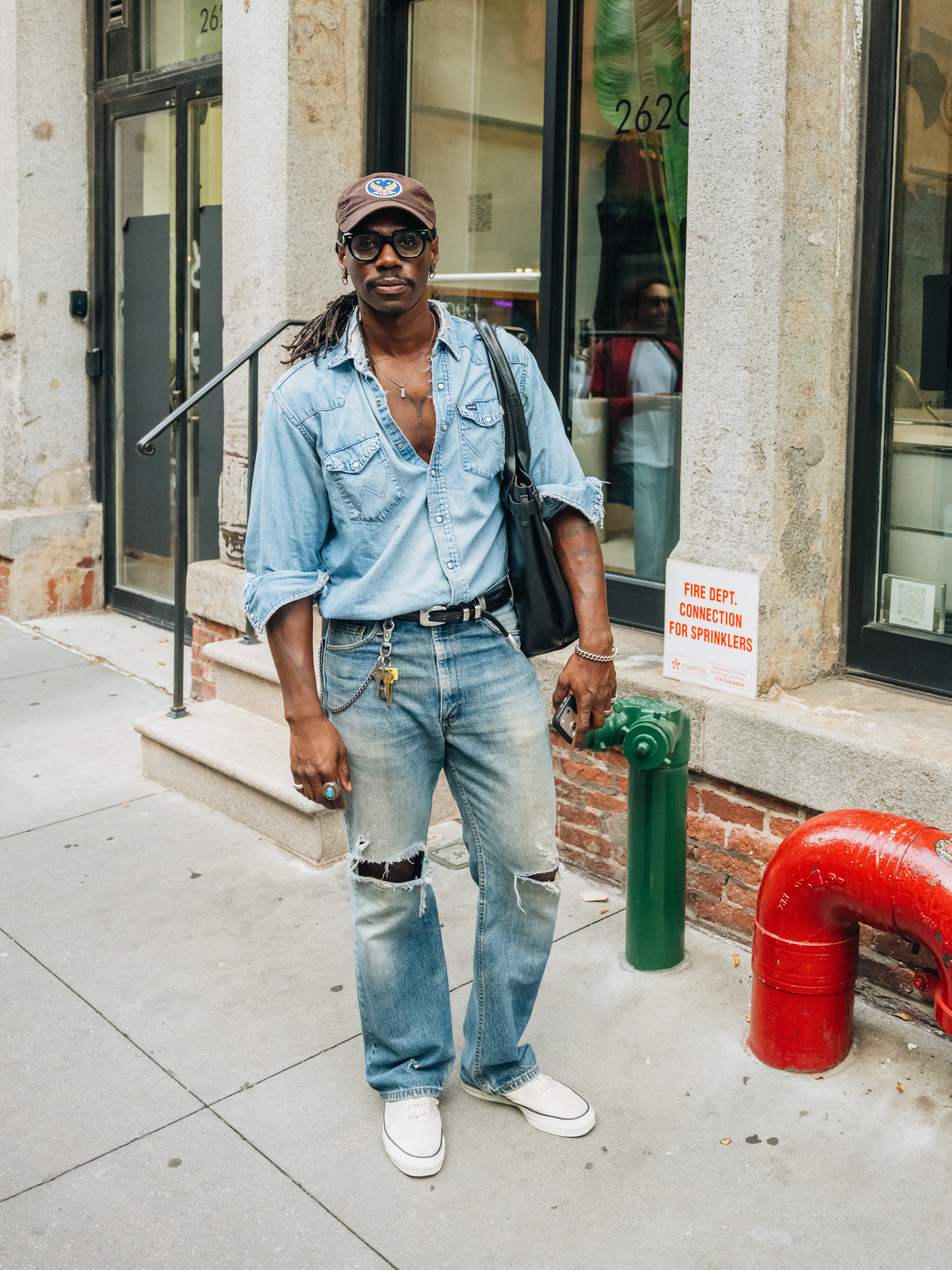 A man with dreadlocks, glasses, and a denim outfit stands on a city sidewalk, holding a phone.