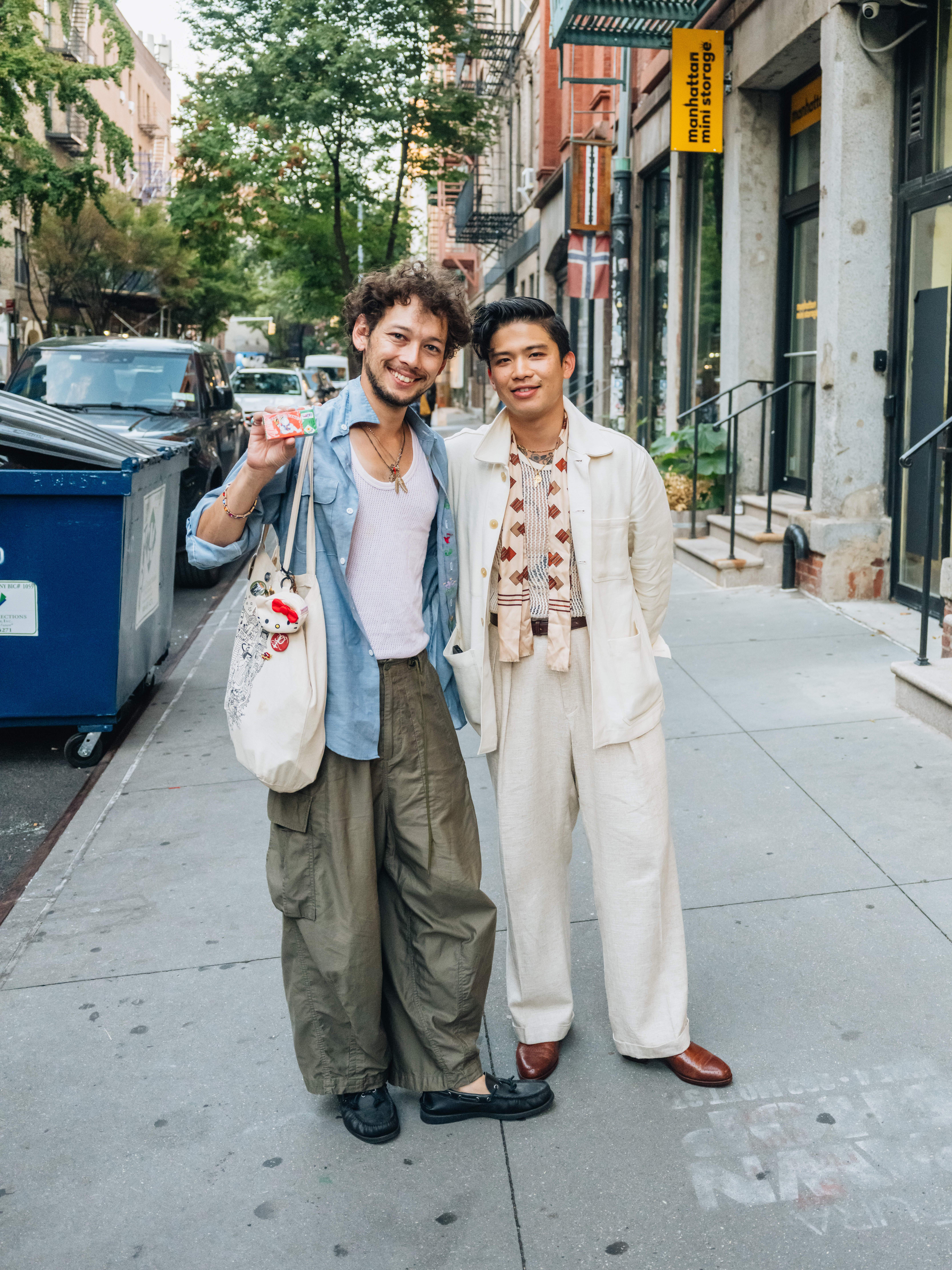 Two men in stylish, loose-fitting clothes smile at the camera on a city sidewalk.