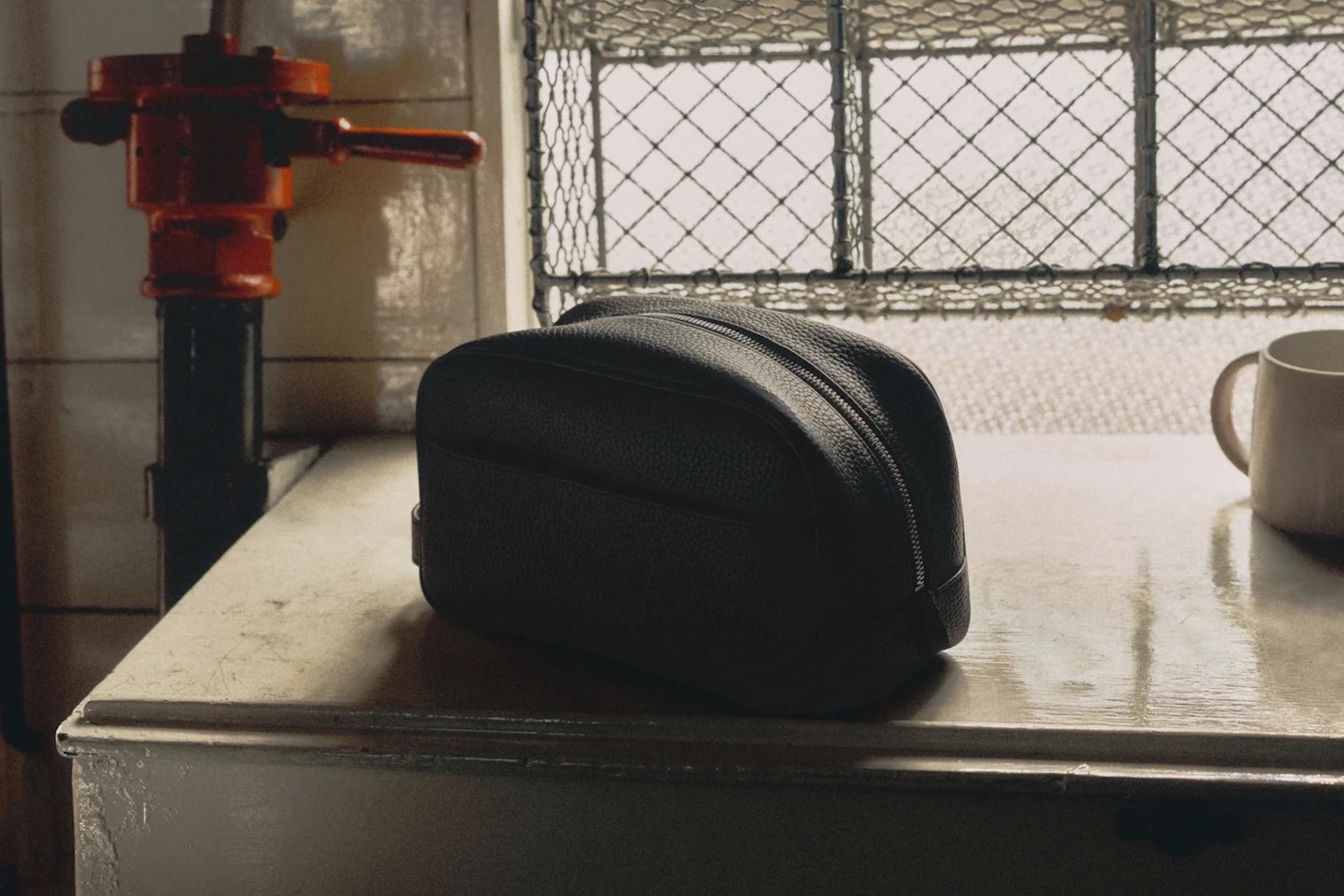Black leather dopp kit on a worn counter, with an orange valve and a white mug in the background.