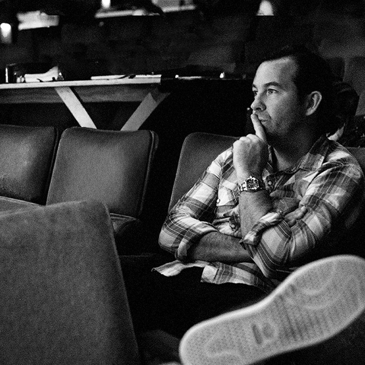 Black and white, Duncan Sheik in a plaid shirt sits in a theater, chin on hand, looking thoughtfully to the right.
