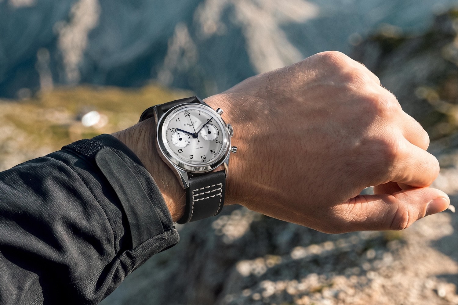 A person's left arm and hand, wearing a silver watch with a black leather band, against a blurred mountain background.