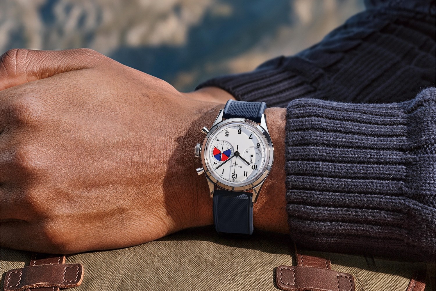 A person's arm wearing a silver watch with a white face, black numbers, and a small red, white, and blue dial.