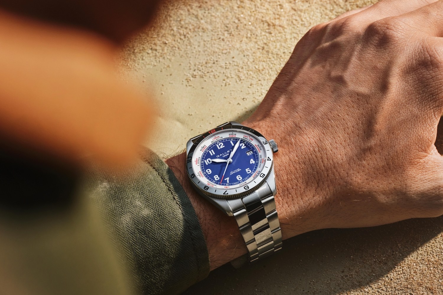 A silver watch with a blue face and white numbers on a metal band, worn on a person's wrist, resting on sand.
