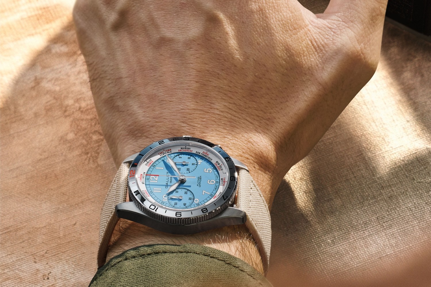 A person's arm wearing a silver watch with a light blue face and a beige strap, against a textured background.
