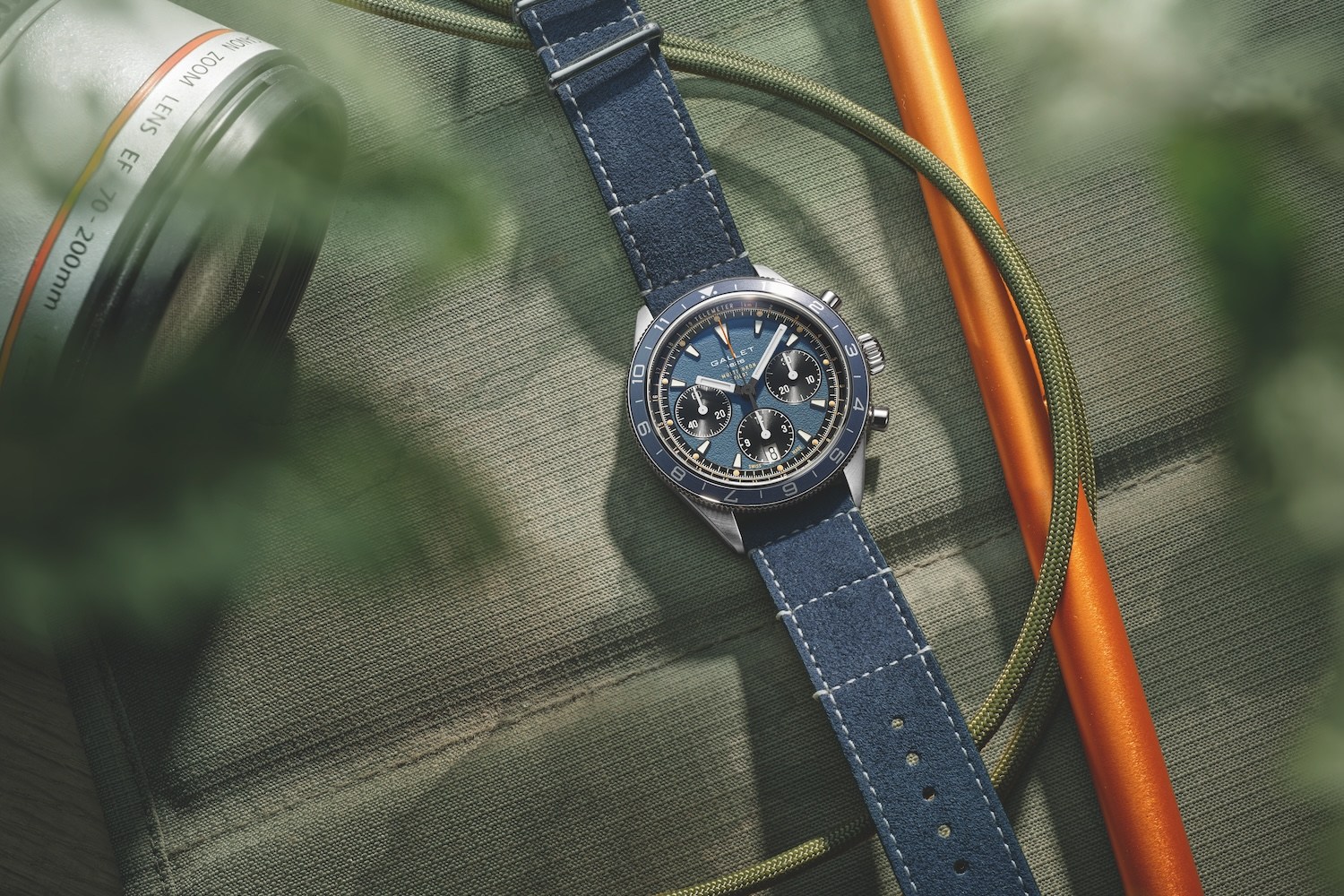 A Gallet watch with a blue face and suede strap, a camera lens, and climbing gear on a green canvas.