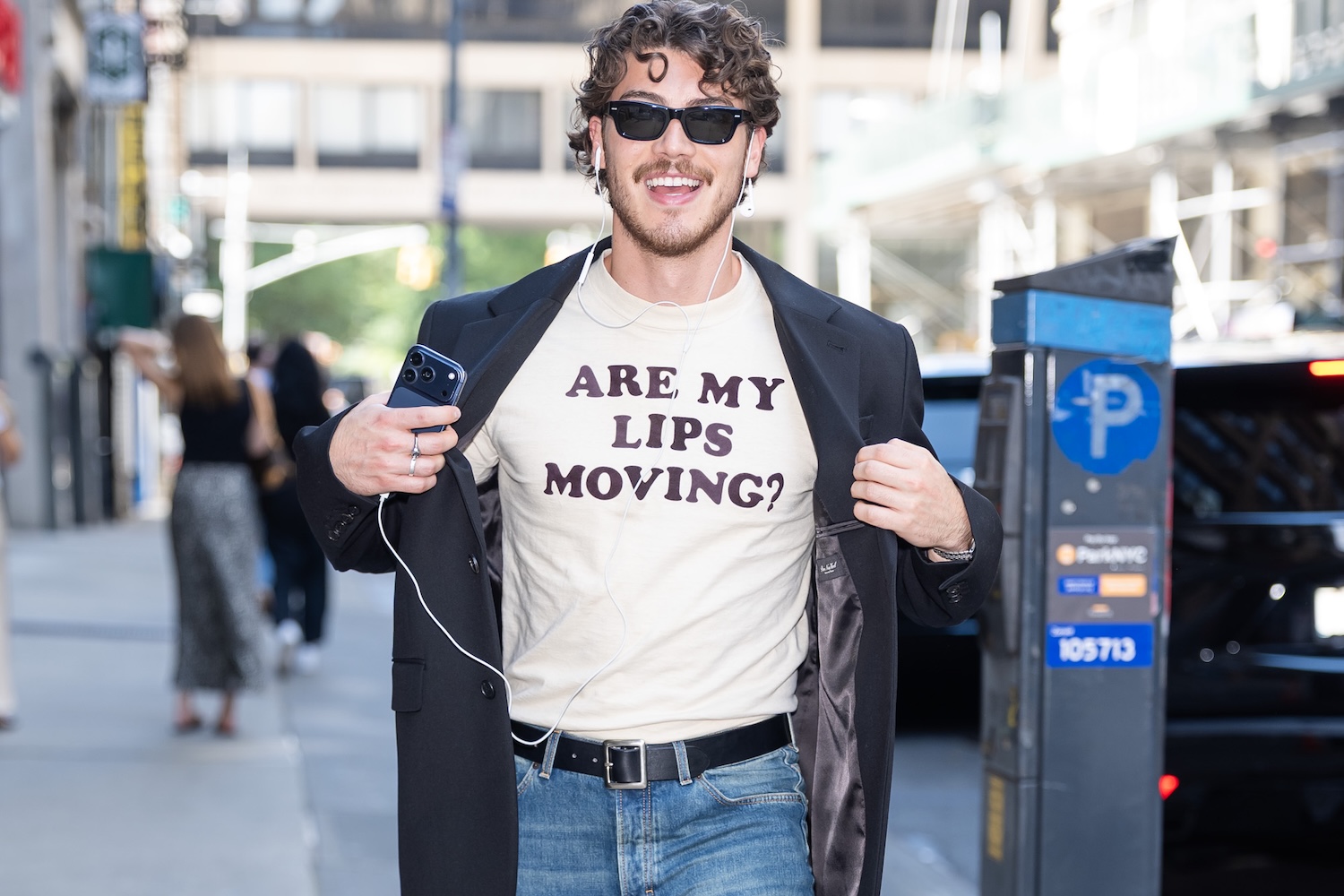 A smiling man in sunglasses and a black blazer over a t-shirt that reads "ARE MY LIPS MOVING?" holds a phone.