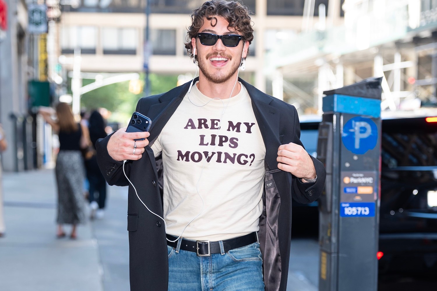 A smiling man in sunglasses and a black blazer over a t-shirt that reads "ARE MY LIPS MOVING?" holds a phone.