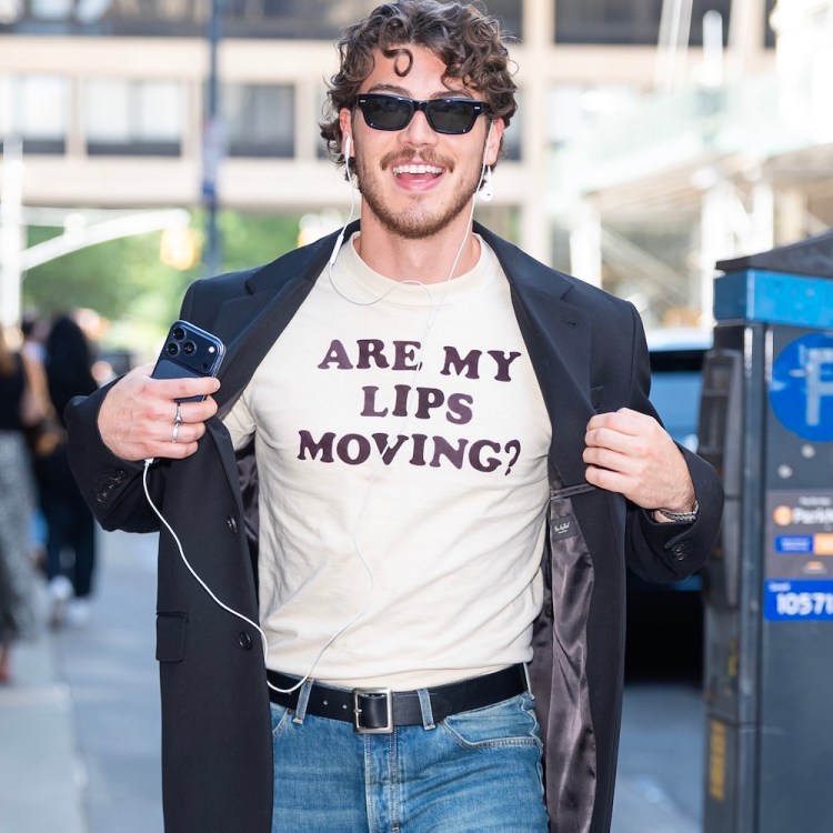 A smiling man in sunglasses and a black blazer over a t-shirt that reads "ARE MY LIPS MOVING?" holds a phone.