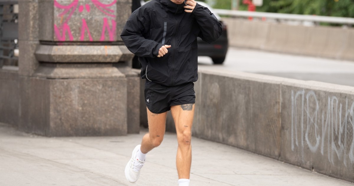 Harry Styles in a black jacket, shorts, and a gray cap runs on a sidewalk next to a concrete barrier.