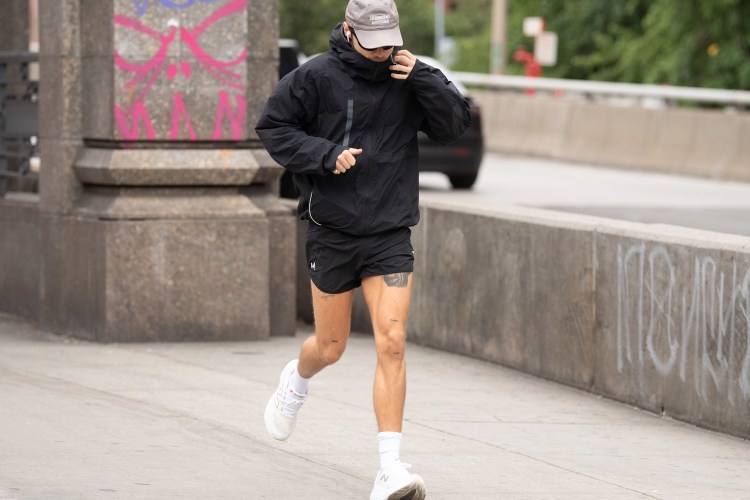 Harry Styles in a black jacket, shorts, and a gray cap runs on a sidewalk next to a concrete barrier.