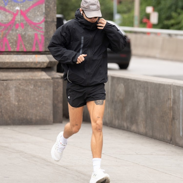 Harry Styles in a black jacket, shorts, and a gray cap runs on a sidewalk next to a concrete barrier.