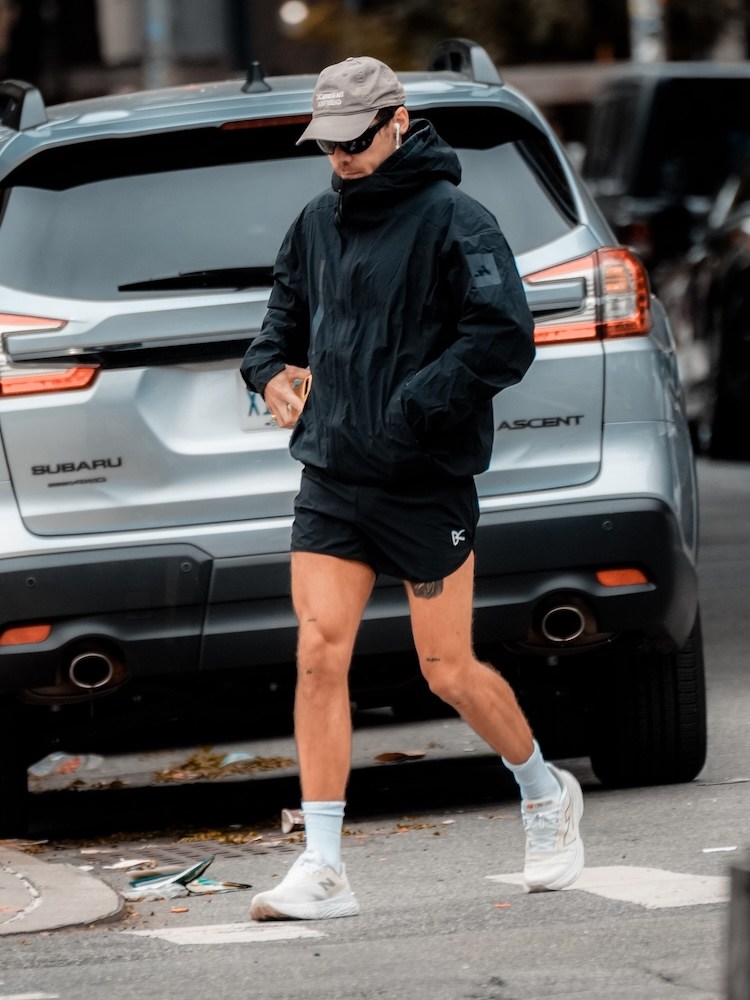 Man in a black jacket, shorts, and white sneakers walking past a gray Subaru SUV.