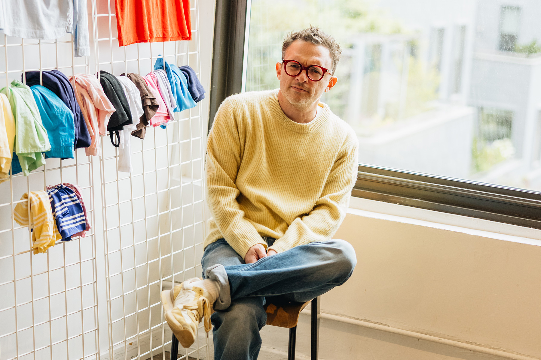 Man in yellow sweater and red glasses sits on a stool, with colorful clothes hanging on a grid behind him.